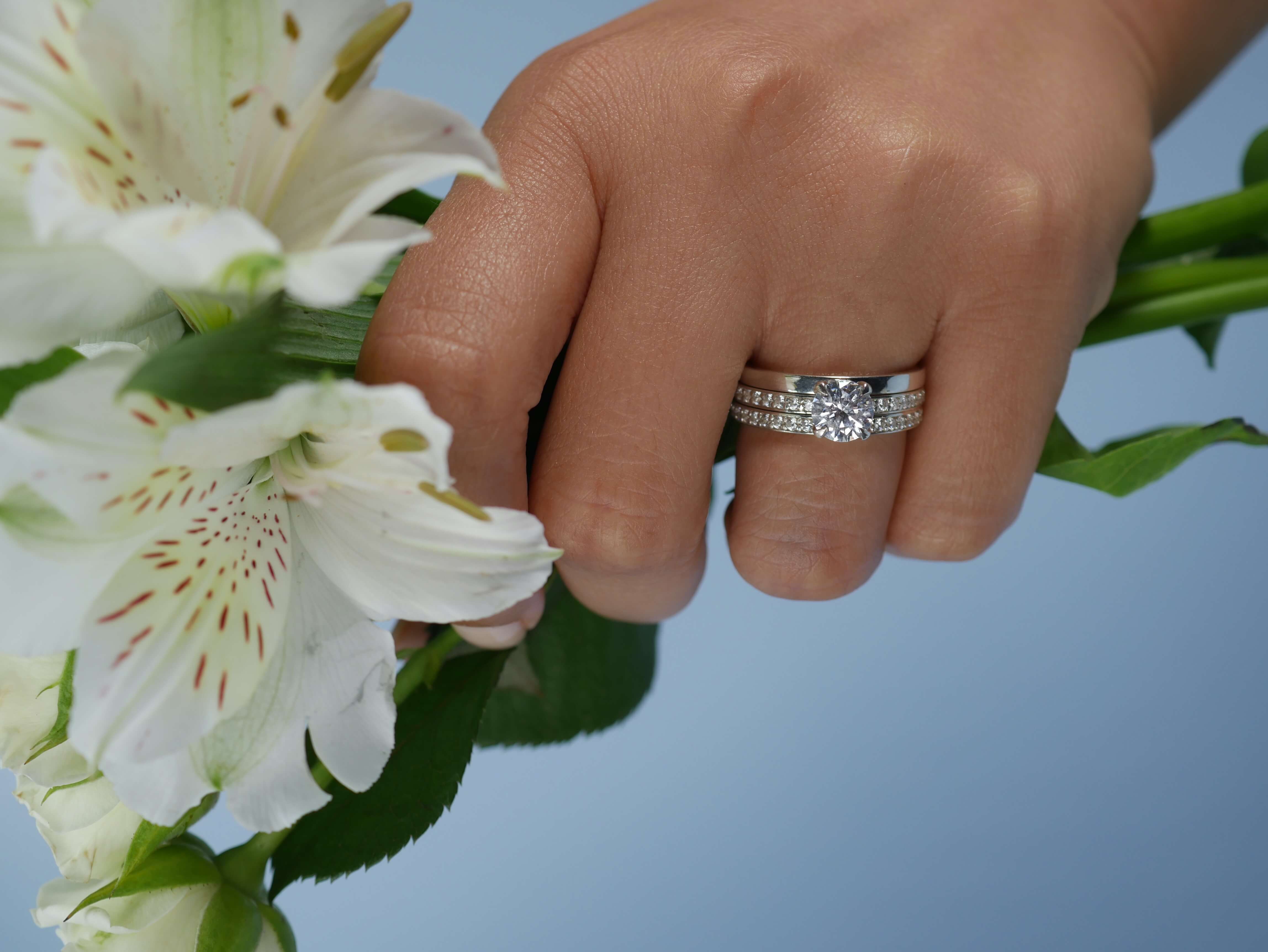 Hand holding a bouquet of white flowers with a diamond engagement ring stacked with a wedding band and diamond eternity band